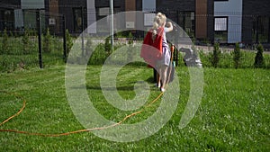 Super girl girl in red cloak mowing lawn in fenced house yard. Corded lawn mower