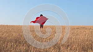Superhero Girl running across field with wheat in red cloak against blue sky.