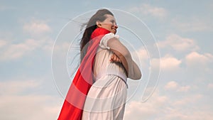 Superhero girl standing on the field in a red cloak, cloak fluttering in the wind. close-up. girl dreams of becoming a