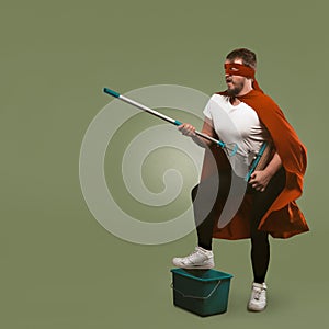 Superhero has fun with cleaning equipment. Man in red cape holds mop and presents himself as musician with guitar