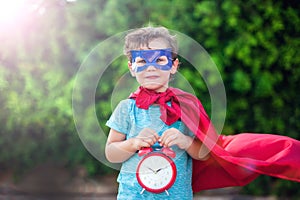 Superhero kid with alarm clock against green background outdoor. Childhood, success and and power concept