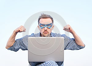 Superhero male in blue mask holds laptop with strength or pumped gesture outdoor