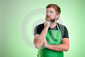 Supermarket employee with green apron and black t-shirt holding hand under his chin, having doubtful