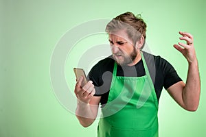 Supermarket employee with green apron and black t-shirt holding smartphone, texting, looking nervously