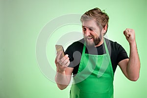 Supermarket employee with green apron and black t-shirt holding smartphone, texting messages happy face