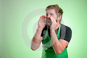 Supermarket employee with green apron and black t-shirt shouting with his hand to his mouth