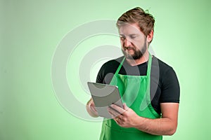 Supermarket employee with green apron and black t-shirt takes notes, looking at the camera