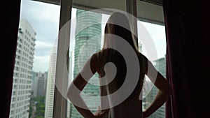 Superslowmotion shot of successful rich young woman standing by the window in her downtown apartment with panoramic view