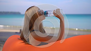 Superslowmotion shot of a young woman on a tropical beach sits on an inflatable sofa and drinks water from a multi