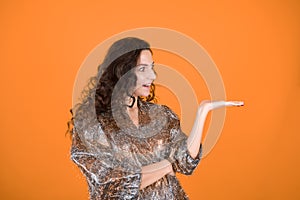 Suprised woman with her palm up over orange background. Caucasian girl in shiny clothes shows something virtual.