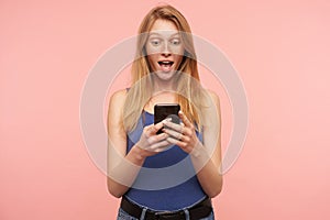 Suprised young pretty long haired redhead woman in blue shirt looking excitedly on screen of her smartphone while reading