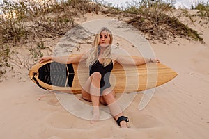 Surf girl in swimsuit sitting with surfboard on beach with warm sunset or sunrise tones