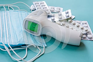 Surface thermometer with digital green colored screen showing high body temperature on pile of medical masks and pills in blisters