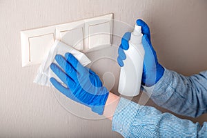 Surfaces disinfection. Houseworker in rubber blue gloves clean light switch with cloth on wall by alcohol spray and rag. New