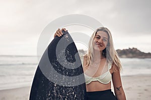 A surfboard is all you need. a young woman standing on the beach with her surfboard.