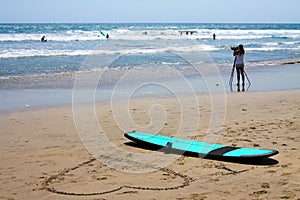 A surfboard is lying on the beach. Next to it, a large heart is drawn in the sand. Good weather, small waves.