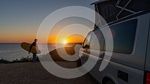 Surfer boy sitting near his mini van and looking on the ocean at summer sunset with a surfboard on her side