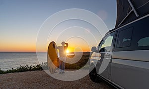 Surfer boy sitting near his mini van and looking on the ocean at summer sunset with a surfboard on her side