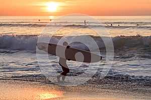 Surfer carrying the surfboard into water with waves in the sunset