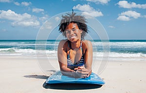 Surfer girl with afro hairstyle smiling, lying down on blue surfboard