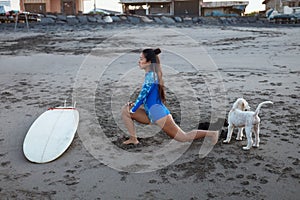Surfer Girl. Surfing Woman Stretching On Sandy Beach. Asian Brunette In Blue Wetsuit With Surfboard And Dog.