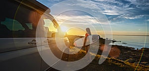 Surfer girl walking near her mini van and looking on the ocean at summer sunset  with a surfboard on her side