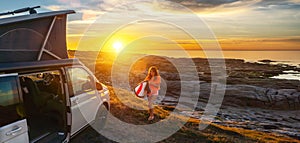 Surfer girl walking near her mini van and looking on the ocean at summer sunset  with a surfboard on her side