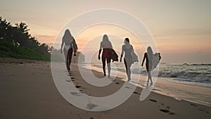 Surfer girls walking along the beach at sunrise. Female surfers carrying surfboards walking on beach along ocean at dawn