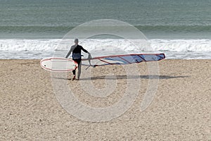 Surfer on a golden sand beach heading into the sea with his windsurf board