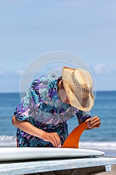 Surfer putting an orange fin into a surfboard in Japan, he is wearing a Hawaiian shirt and straw hat.