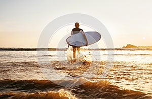 Surfer runs with surfboard towards ocean waves ta sunset time