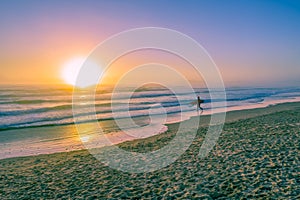 Surfer silhouette walking on ocean beach at sunset.