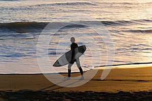 Surfer in silhouette walks to water`s edge carrying surfboard back-lit by golden light of setting sun