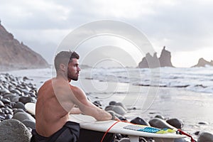Surfer sitting at the beach with his surfboard, athletic guy doing extreme sport, photo with copy space