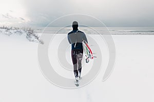 Surfer in wetsuit with surfboard run to ocean. Snowy day with male surfer