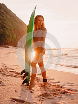 Surfer woman in swimwear hold surfboard at tropical beach with sunset tones