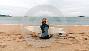 Surfer woman with wetsuit and surfboard sitting on the sand looking aside on the beach