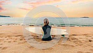 Surfer woman with wetsuit and surfboard sitting on the sand looking at the sea