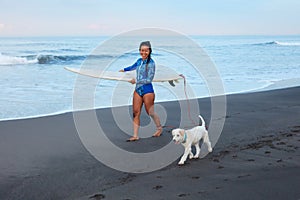 Surfing Girl. Beautiful Woman Walking With Dog On Ocean Beach. Brunette In Blue Wetsuit Carrying Surfboard.