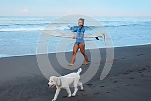 Surfing Girl. Beautiful Woman Walking With Dog On Sandy Beach. Smiling Brunette In Blue Wetsuit Carrying Surfboard.