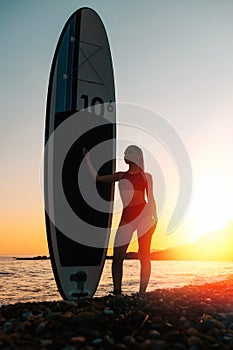 Surfing. Silhouette of a young slender woman holding a sup board. In the background, the ocean and the sunset. Summer vacation at