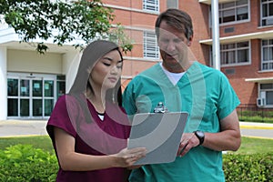 Surgeon or Doctor or Physician or Clinician and Asian Nurse Stand in Front of Hospital Discussing Patient Chart