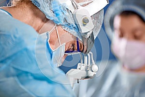 Surgeon and his assistant performing cosmetic surgery in hospital operating room. Surgeon in mask wearing loupes during