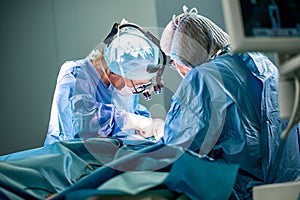 Surgeon and his assistant performing cosmetic surgery in hospital operating room. Surgeon in mask wearing loupes during