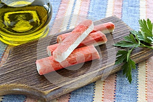 Surimi crab sticks and parsley on a brown cutting board near jar with olive oil. Seafood and ingredient for salads. Healthy eating