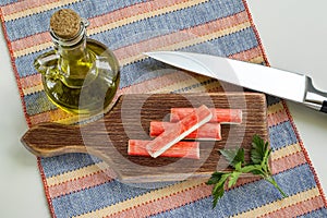 Surimi crab sticks and parsley on a brown cutting board near jar with olive oil and sharp chef knife. Seafood and ingredient for