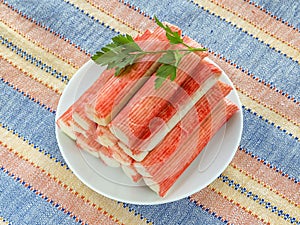 Surimi crab sticks and parsley on a white saucer over a striped table mat. Seafood and ingredient for salads. Healthy eating and