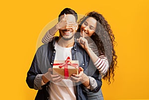 Surprise Present. Romantic young arab woman covering boyfriend's eyes and giving gift