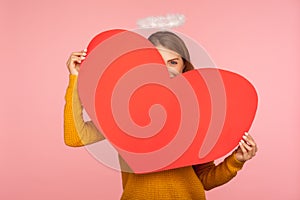 Surprise on Valentine`s day. Portrait of funny girl with halo over head hiding behind big red heart and looking playfully