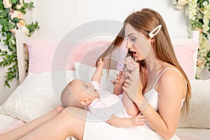 Surprise young beautiful mother holding her daughter 6 months on her lap on a white bed and looking at her open mouth, mother`s